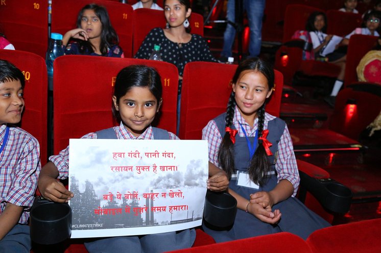 Two girls in a cinema-seated auditorium holding up a hand-lettered Hindi poster about air and water pollution