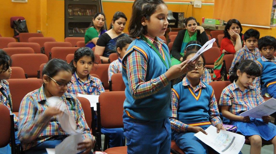 A girl standing to read from a worksheet in a school auditorium while classmates follow on their own papers, three facilitators at the back