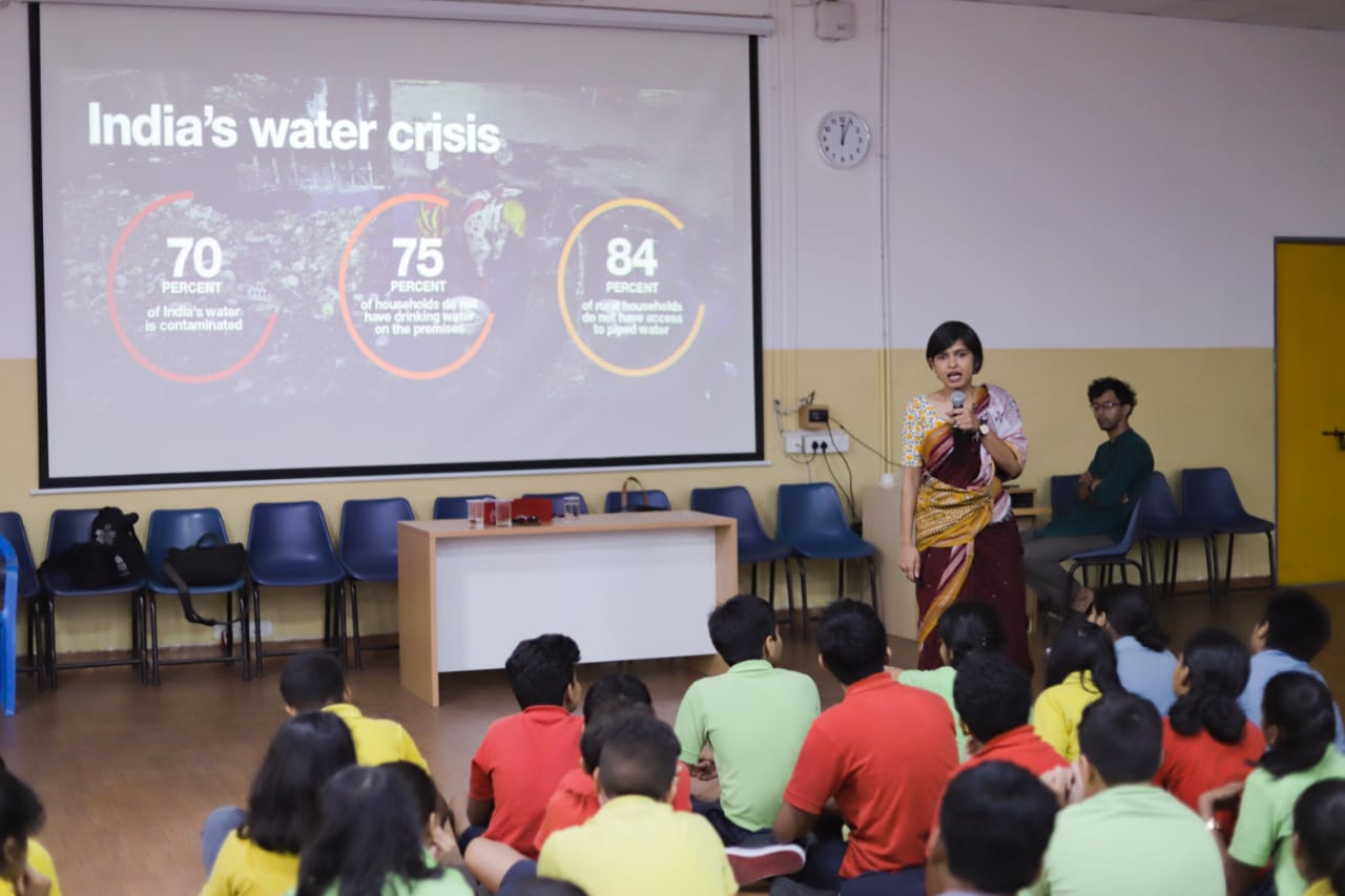 A speaker addressing seated schoolchildren in a hall, the screen behind reading India’s water crisis with three figures on it