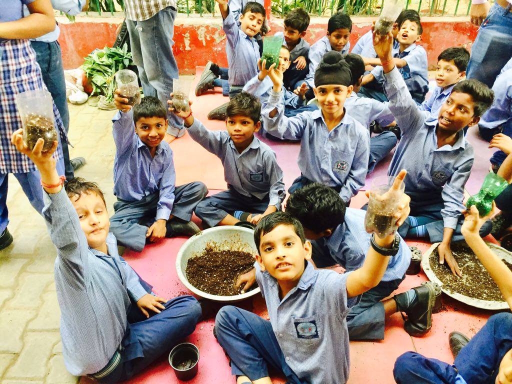 Children in school uniform sitting cross-legged on a mat around two bowls of compost, several holding up a fistful of soil