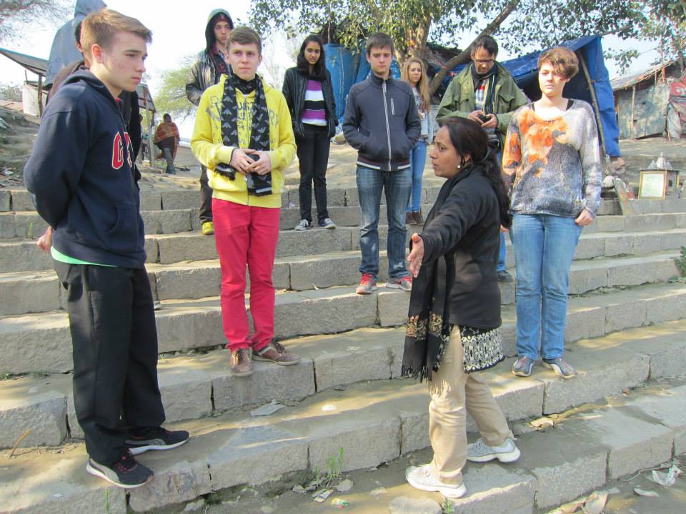 Teenagers standing on stone ghat steps above an informal riverside settlement while a facilitator addresses them