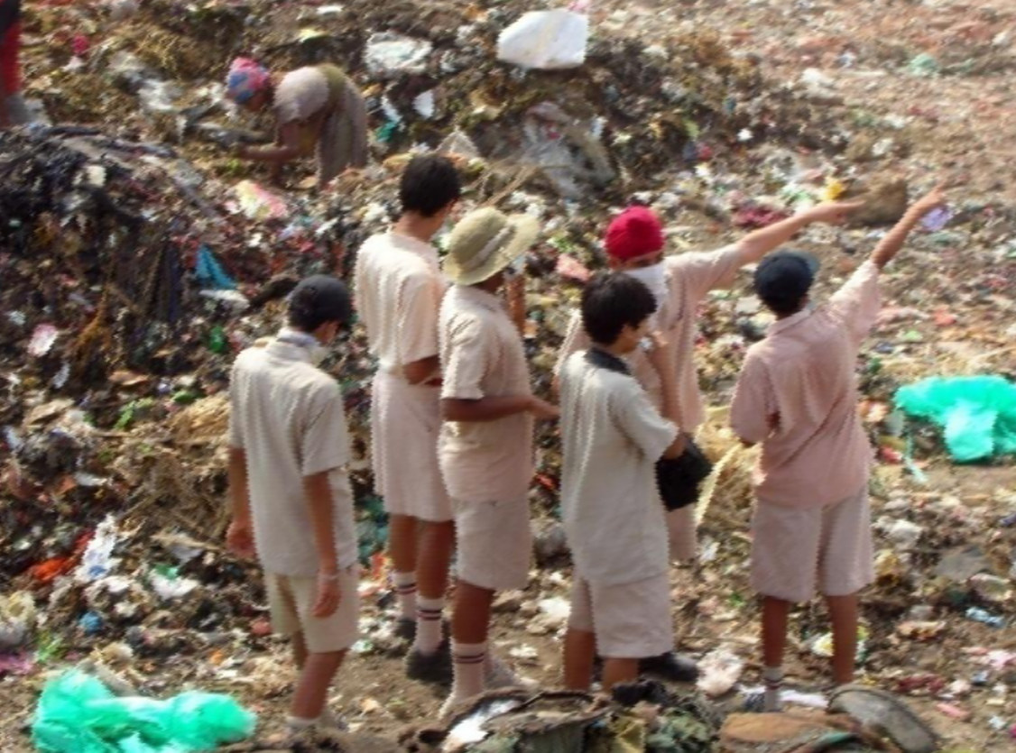 Children in beige school uniforms standing at the edge of a landfill mound, a rag-picker working the waste behind them