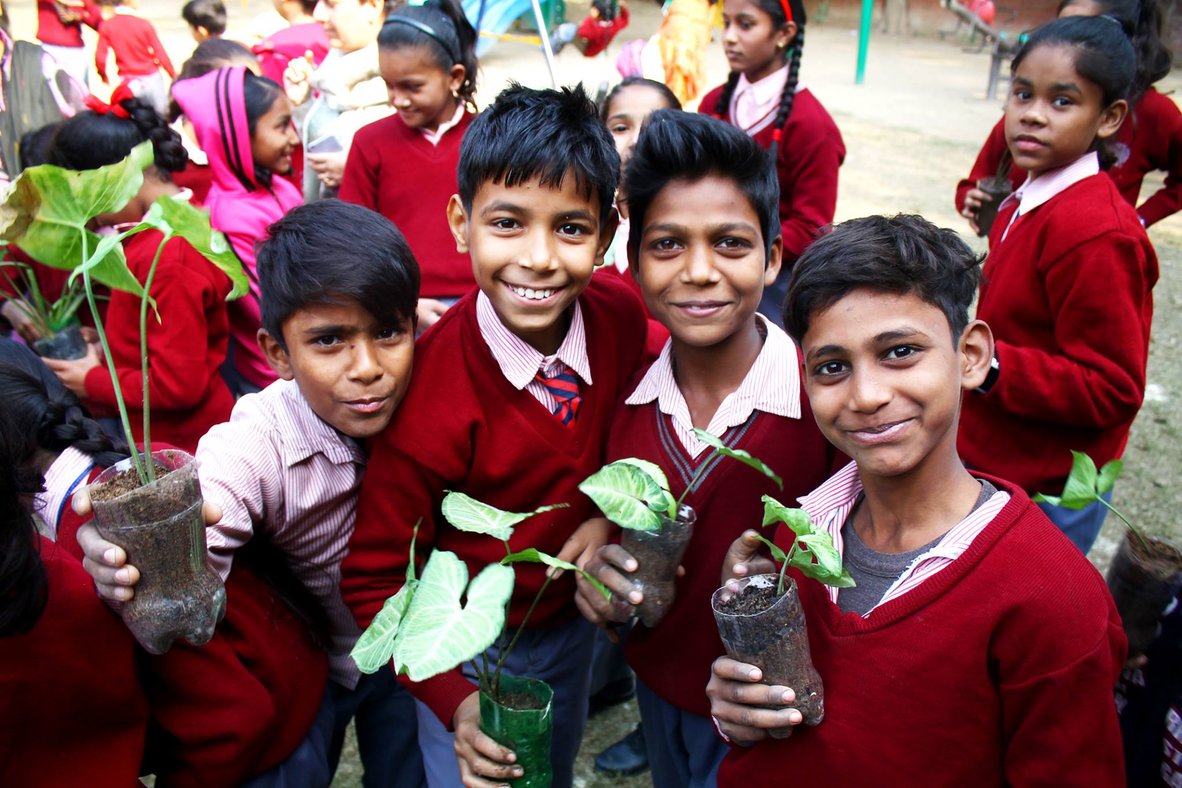 A group of children in maroon school uniforms, each holding up a sapling growing in a repurposed plastic cup