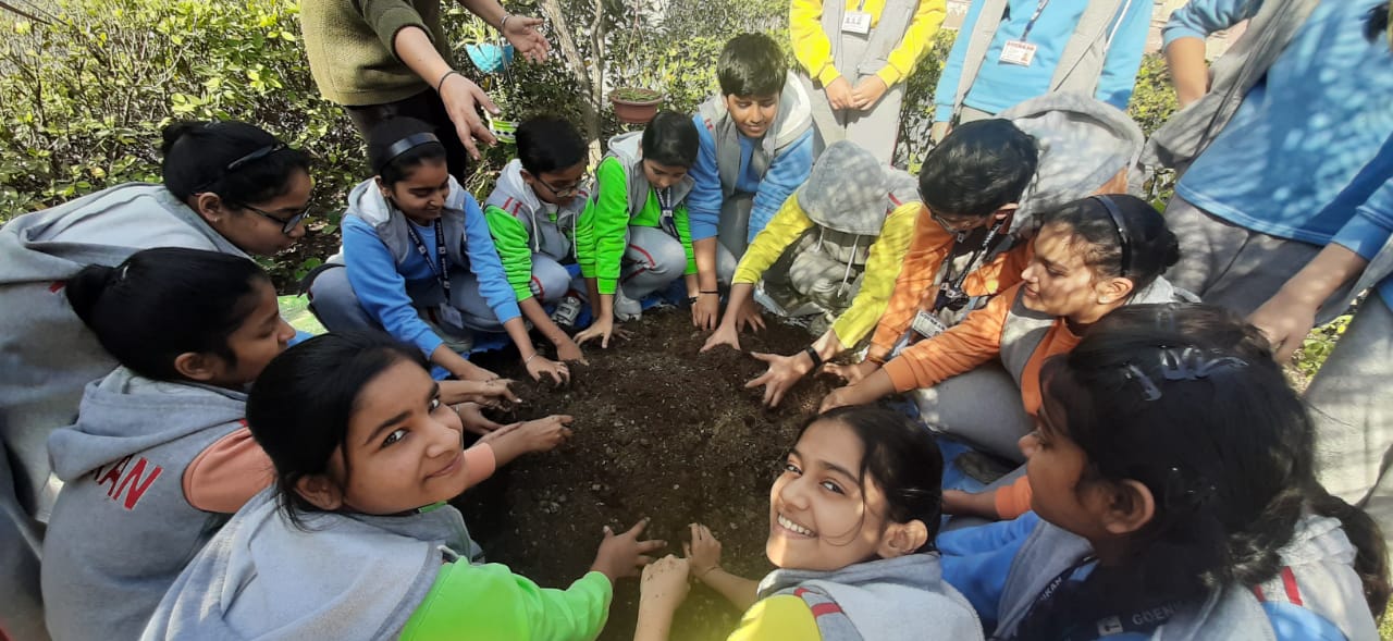 A tight circle of students in school jackets, all hands together packing soil around a newly planted tree