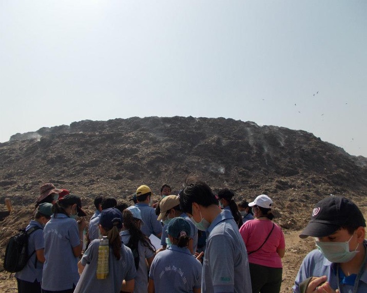 A group of masked students standing at the foot of a large dark landfill mound under a hazy sky