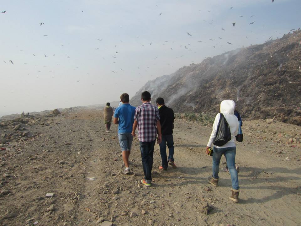 A small group walking a dirt track beside a smoking landfill, birds circling above it