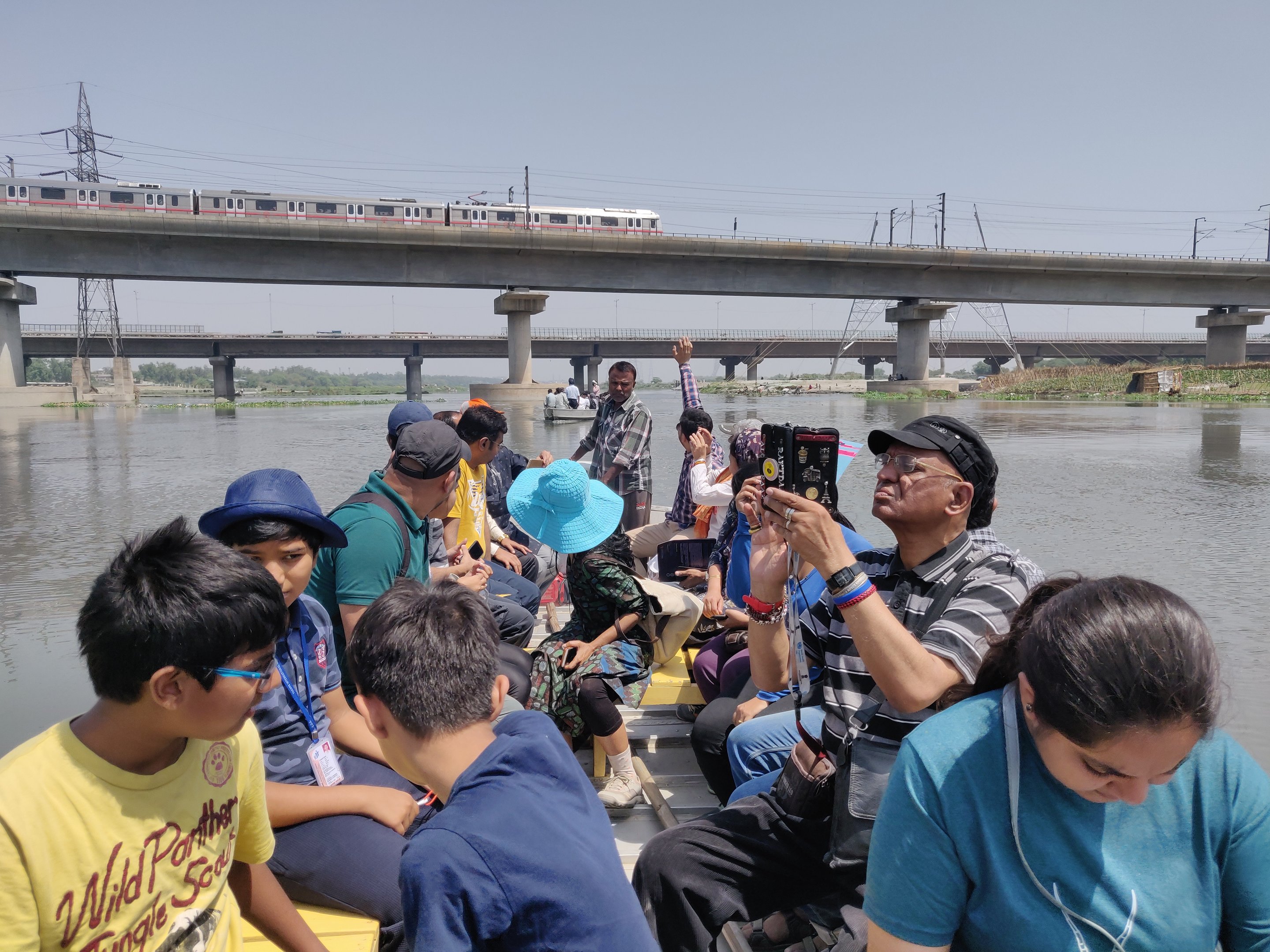A group seated in a boat on the Yamuna, a Delhi Metro train crossing the bridge overhead