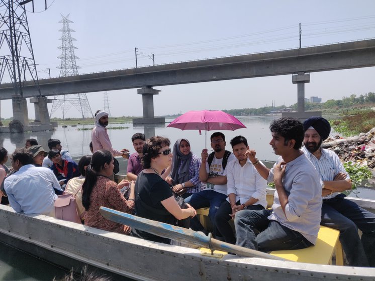 A group seated in a boat on the Yamuna under a pink umbrella, a road bridge behind them