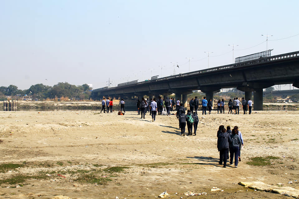 A line of students walking across a dry, cracked stretch of Yamuna riverbed towards a road bridge