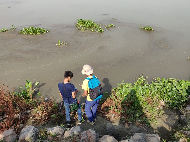 Two people standing on the rocky edge of the Yamuna looking out over water matted with hyacinth