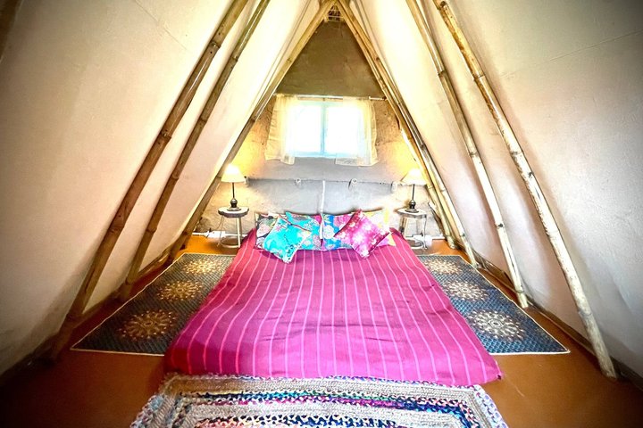 Inside an A-frame mud house — a bed under the bamboo apex, a small window at the far end