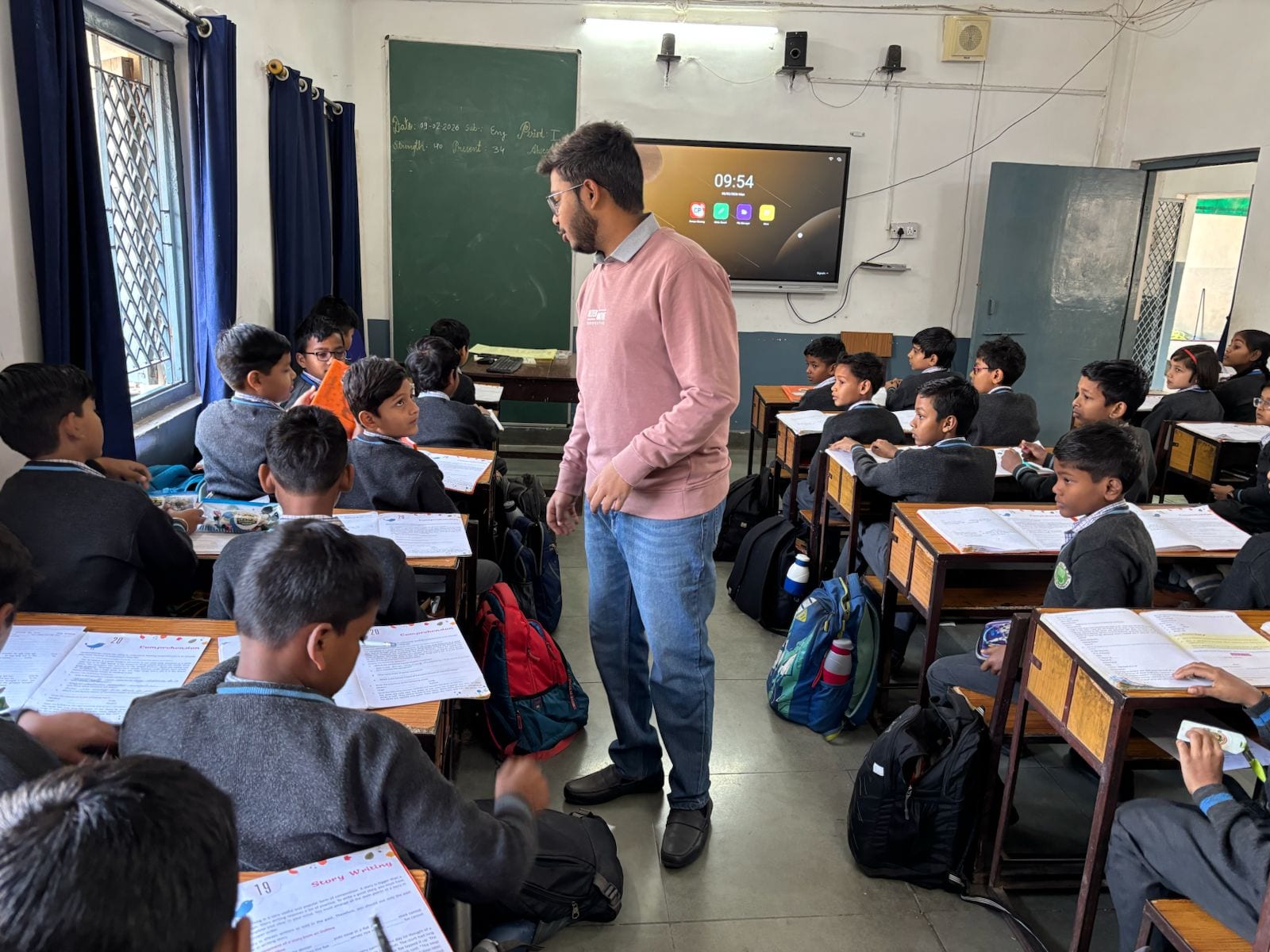 A man in a rose sweater standing in the aisle of a crowded classroom, children in grey school jumpers at wooden desks turning from their open exercise books to look at him, a green chalkboard and a wall-mounted screen behind