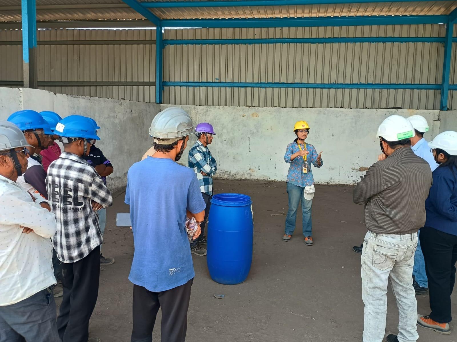 A woman in a yellow hard hat briefing a ring of campus workers in blue, white and purple helmets under an open shed, a blue composting drum standing on the floor between them