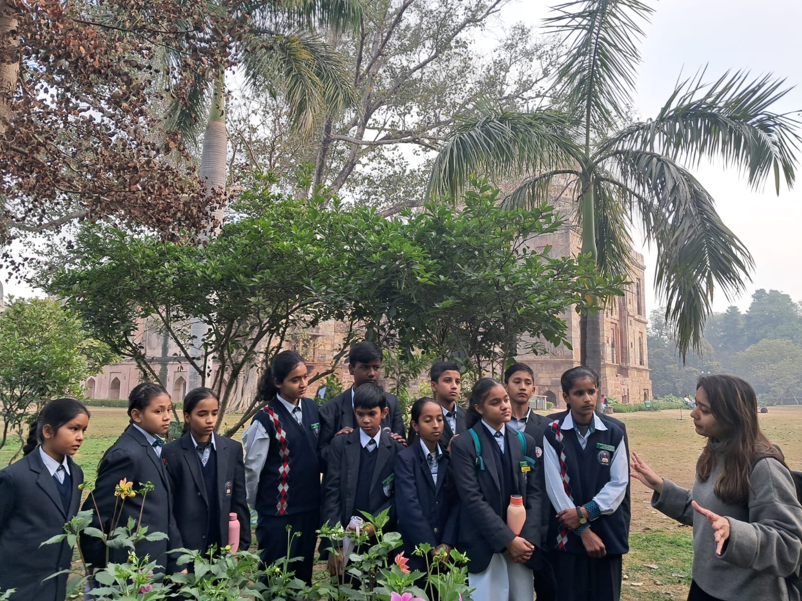 Twelve schoolchildren in grey blazers and striped ties standing on a Lodhi Garden lawn, a facilitator on the right talking to them with both hands raised, a sandstone Lodhi-era tomb and date palms behind them and flowering beds in the foreground