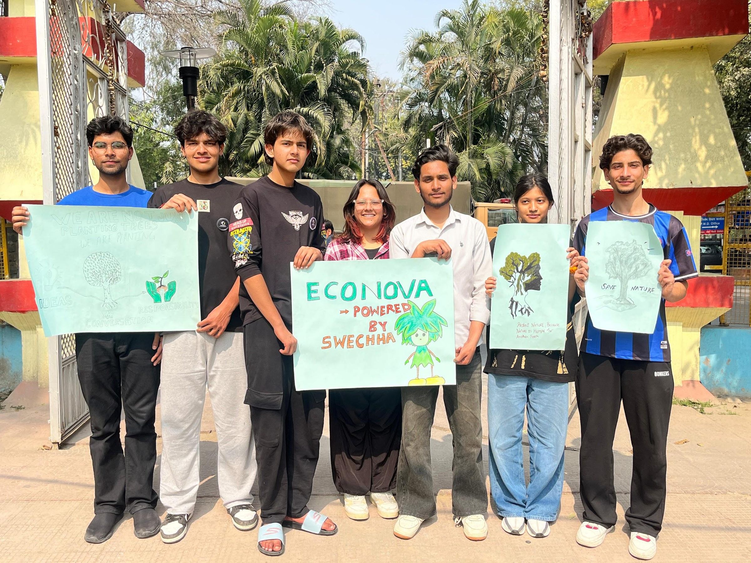 Seven young people standing at a park gate holding hand-painted posters, the middle one lettered ECONOVA, powered by Swechha