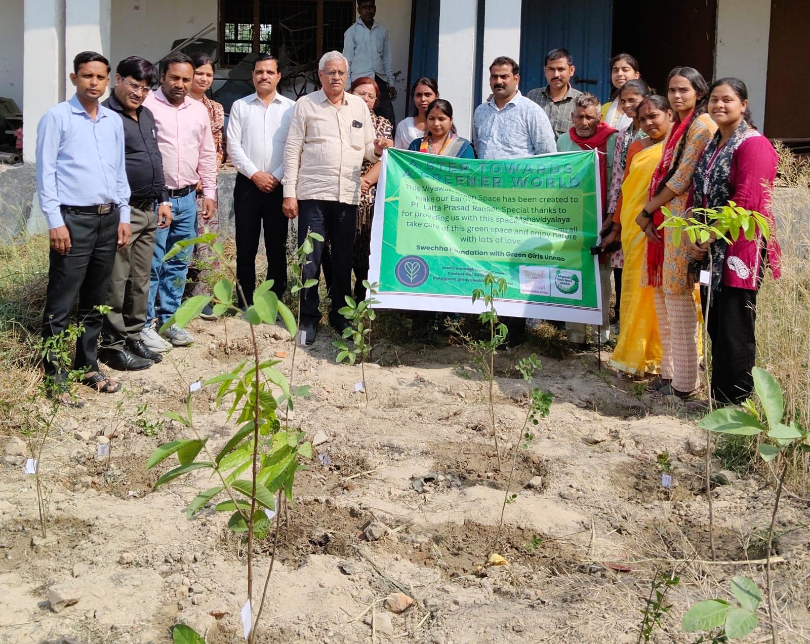 Men and women standing shoulder to shoulder behind a green banner reading A step towards a greener world, rows of labelled knee-high saplings planted in bare stony earth in front of them