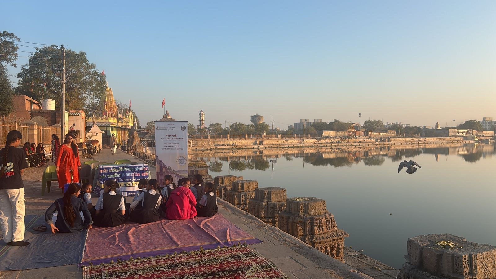 Schoolgirls seated on rugs on the paved edge of Munsar Lake in low morning light, facing a Varso Jal Sanchay banner, with carved stone blocks, a flagged temple and the far bank reflected in still water