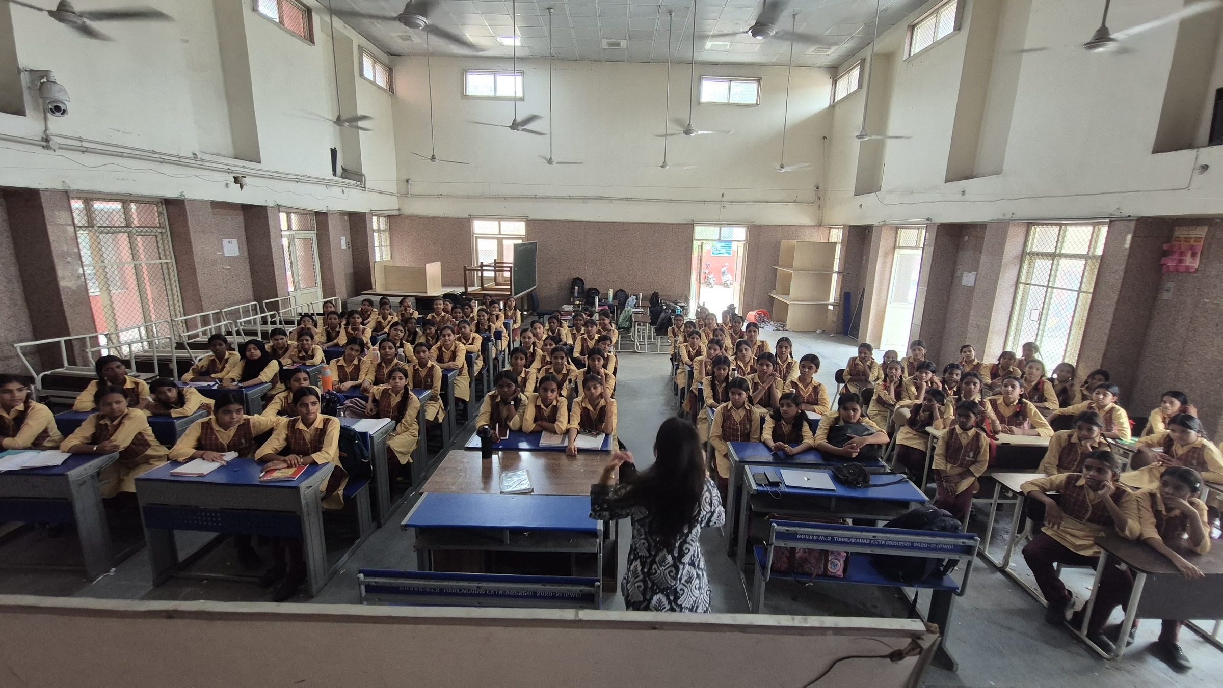 About a hundred students in yellow uniforms seated at desks in a school hall, facing a facilitator standing at a table at the front