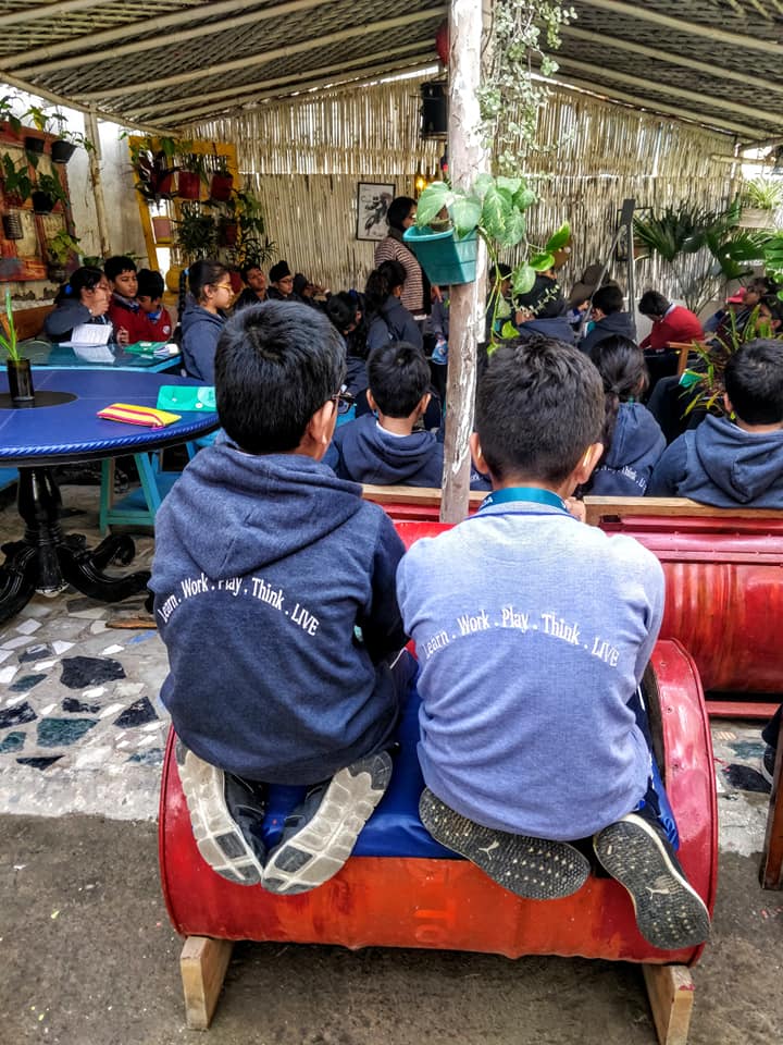 Children seated on a red bench under a bamboo-and-thatch pavilion roof, potted plants hanging above an open-air classroom