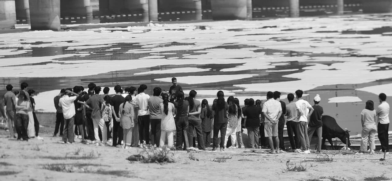 Students standing along the bank facing the foam-covered Yamuna